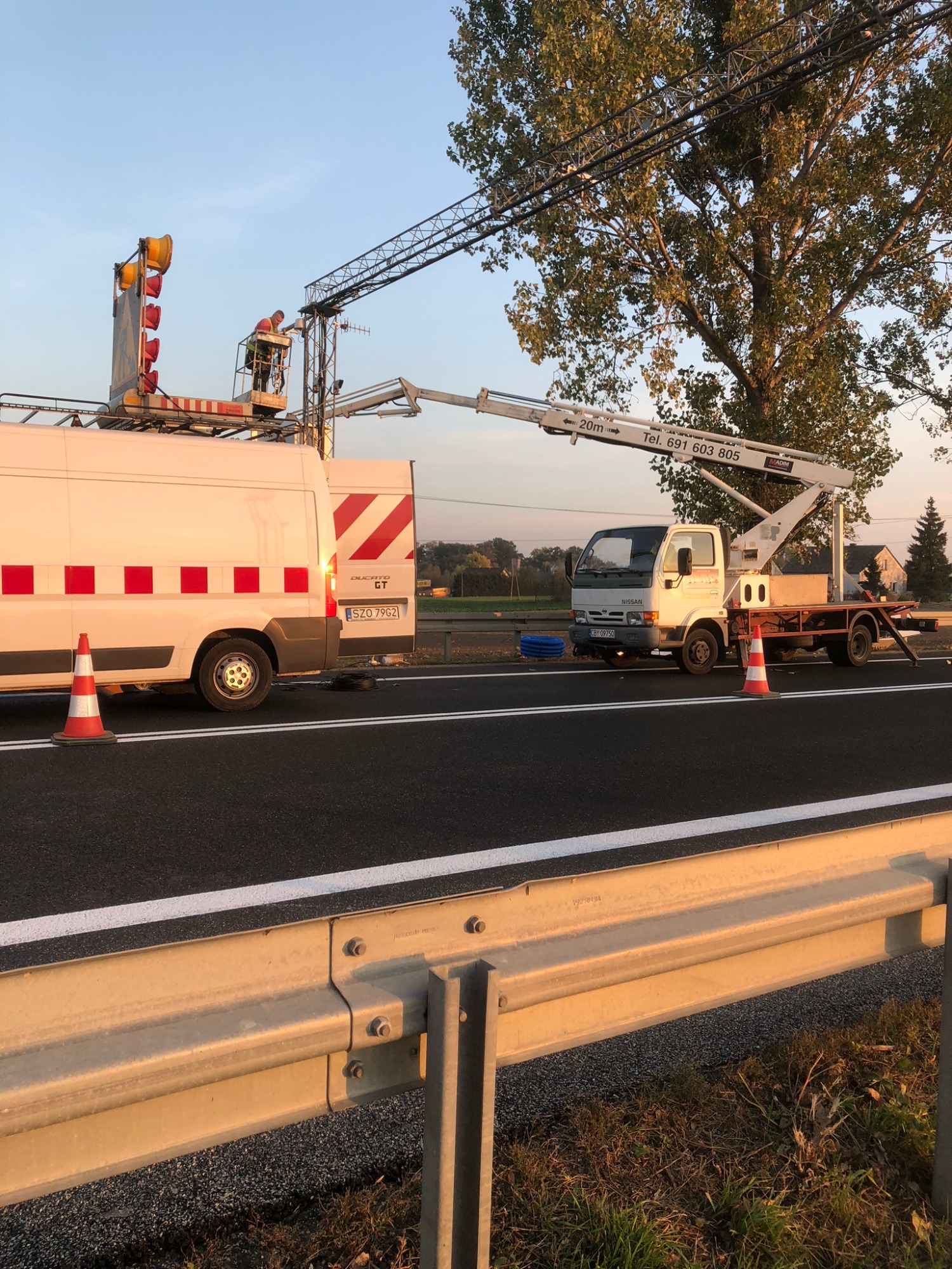 Pracownik w pomarańczowej kamizelce na platformie roboczej na wysokości, przy montażu lub konserwacji oświetlenia drogowego, z użyciem dwóch pojazdów z wysięgnikami na tle drzew i błękitnego nieba.