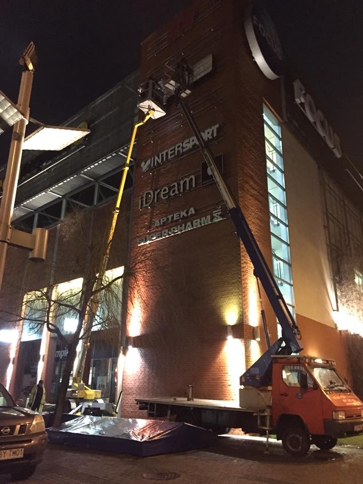 Montaż reklam na elewacji budynku centrum handlowego przy użyciu dwóch wysięgników koszowych, widoczne marki sklepów: Intersport, iDream, Apteka Superpharm.