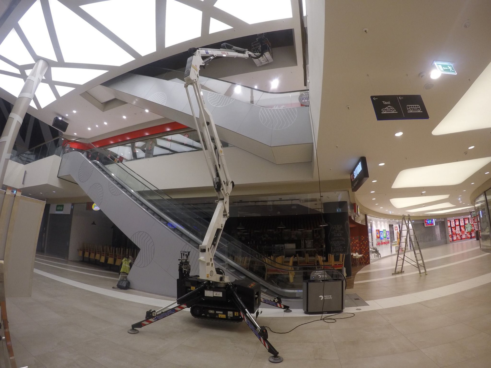 Wnętrze centrum handlowego z rozłożonym białym podnośnikiem na gąsienicach, pracownik w żółtej kamizelce czyszczący podłogę w tle, schody ruchome i sufit z geometrycznym oświetleniem.