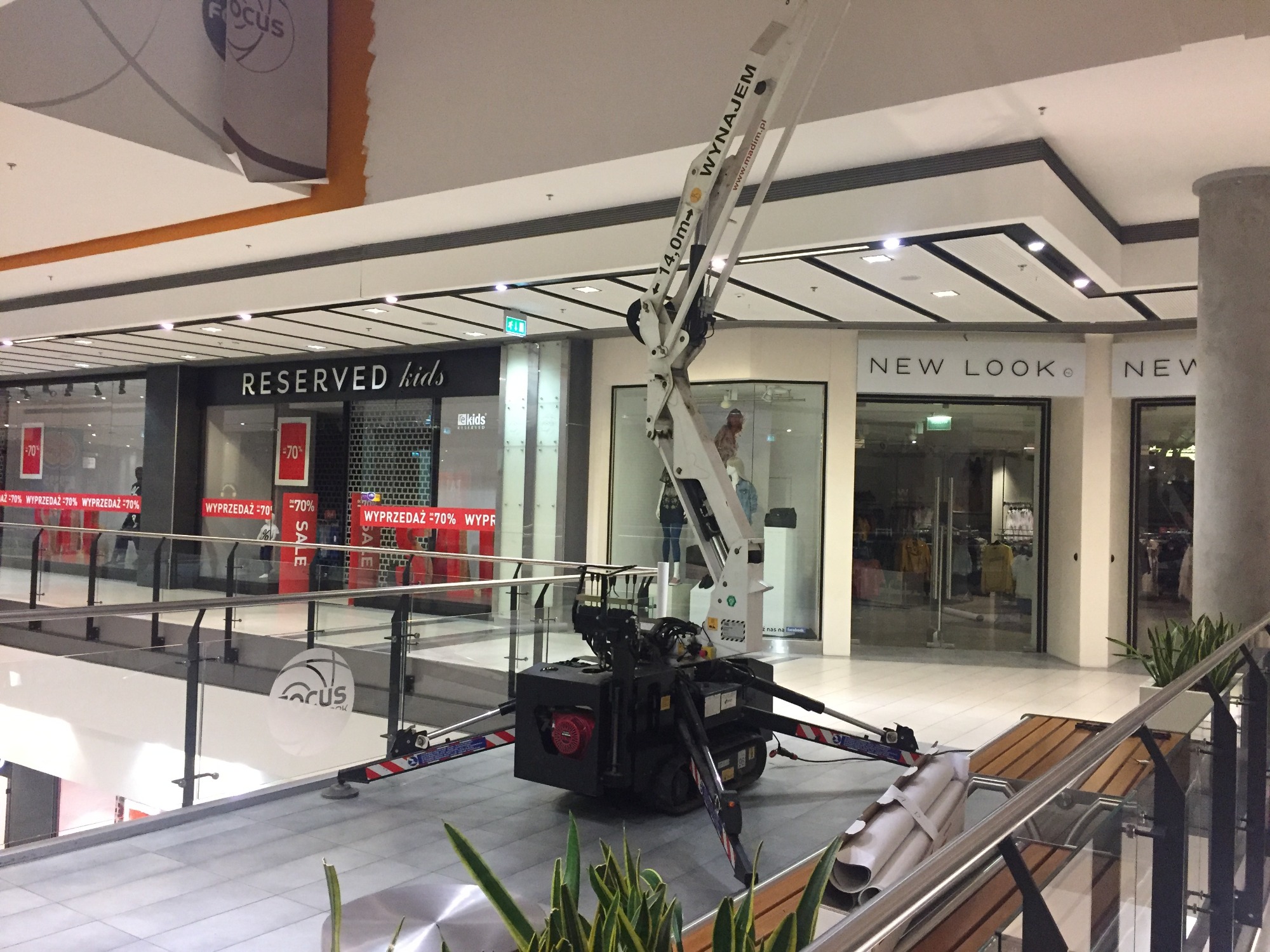 Kompaktowy podnośnik koszowy z wysięgnikiem 14m ustawiony wewnątrz galerii handlowej, widoczne sklepy Reserved kids i New Look w tle.