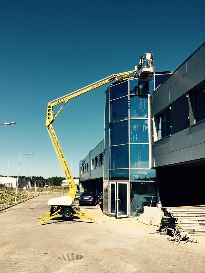Żółty wysięgnik z platformą roboczą przy szklanej fasadzie budynku biurowego, widoczny pracownik w koszu, błękitne niebo w tle.