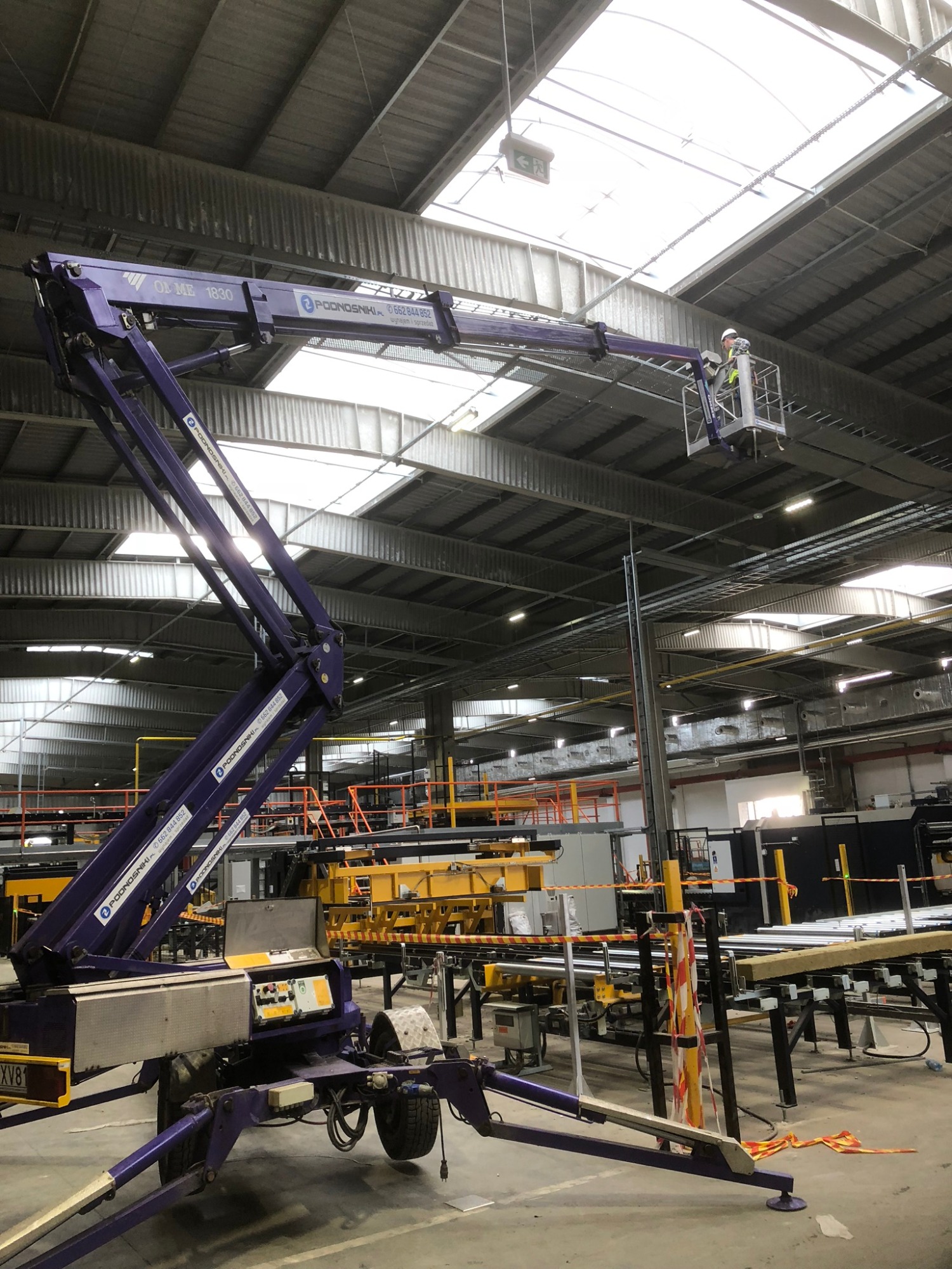 Praca w hali produkcyjnej w Łochowie z wykorzystaniem podnośnika teleskopowego, operator w koszu na wysokości, widoczne świetliki w dachu hali.