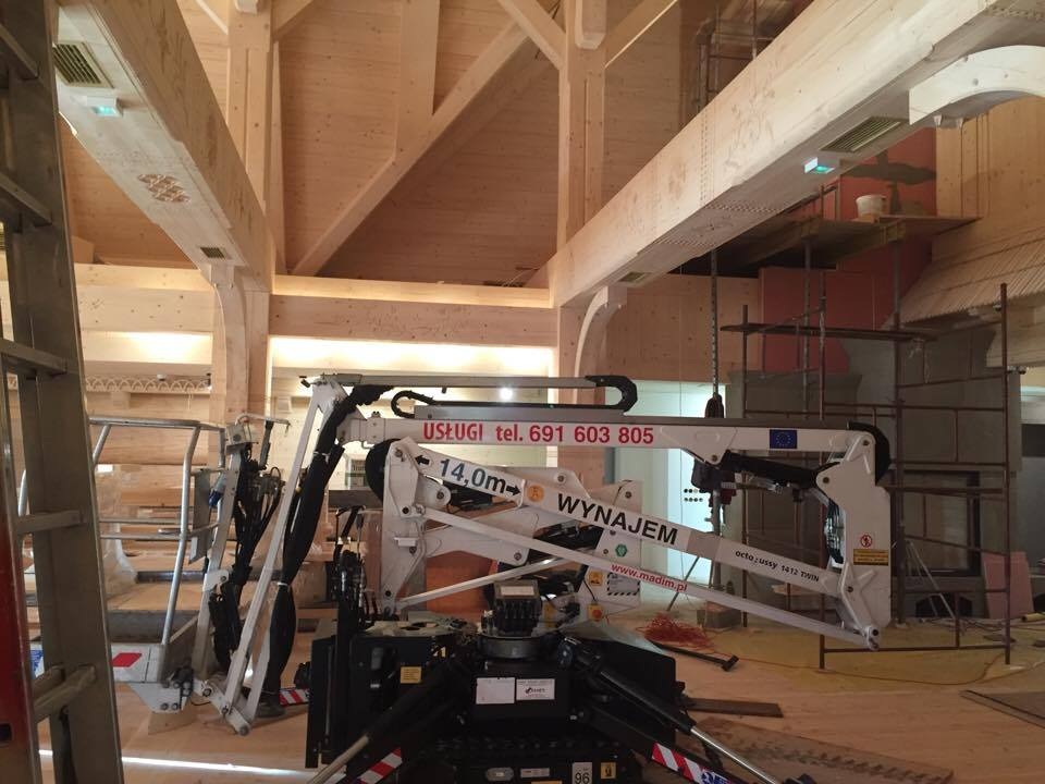Biały sprzęt z wysięgnikiem z napisem 'WYNAJEM' wewnątrz drewnianej konstrukcji budynku w trakcie budowy, widoczne rusztowania i drewniane elementy konstrukcyjne.