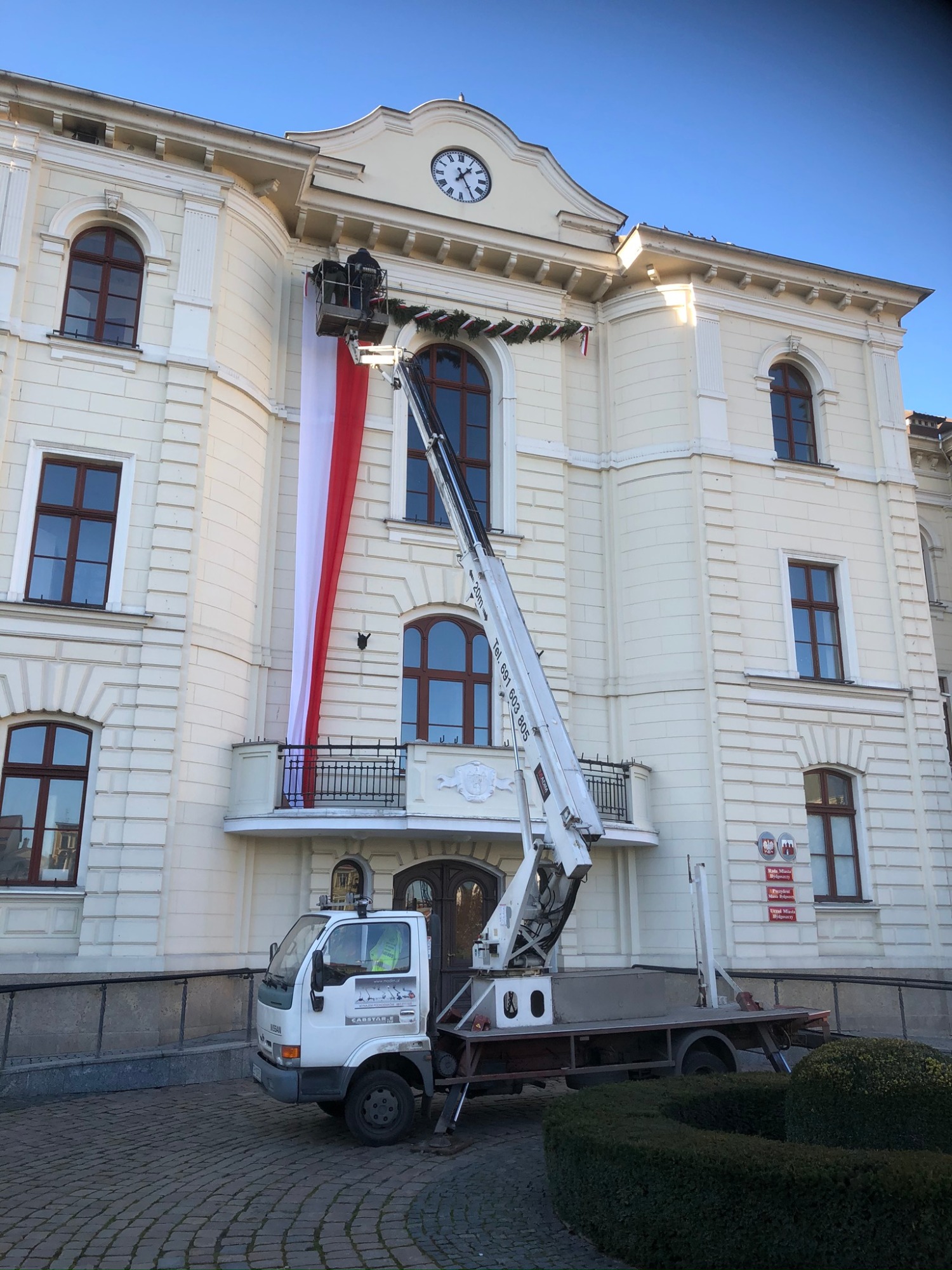 Montaż dekoracji świątecznych na budynku urzędu za pomocą podnośnika koszowego, widoczna flaga Polski i pracownik w koszu