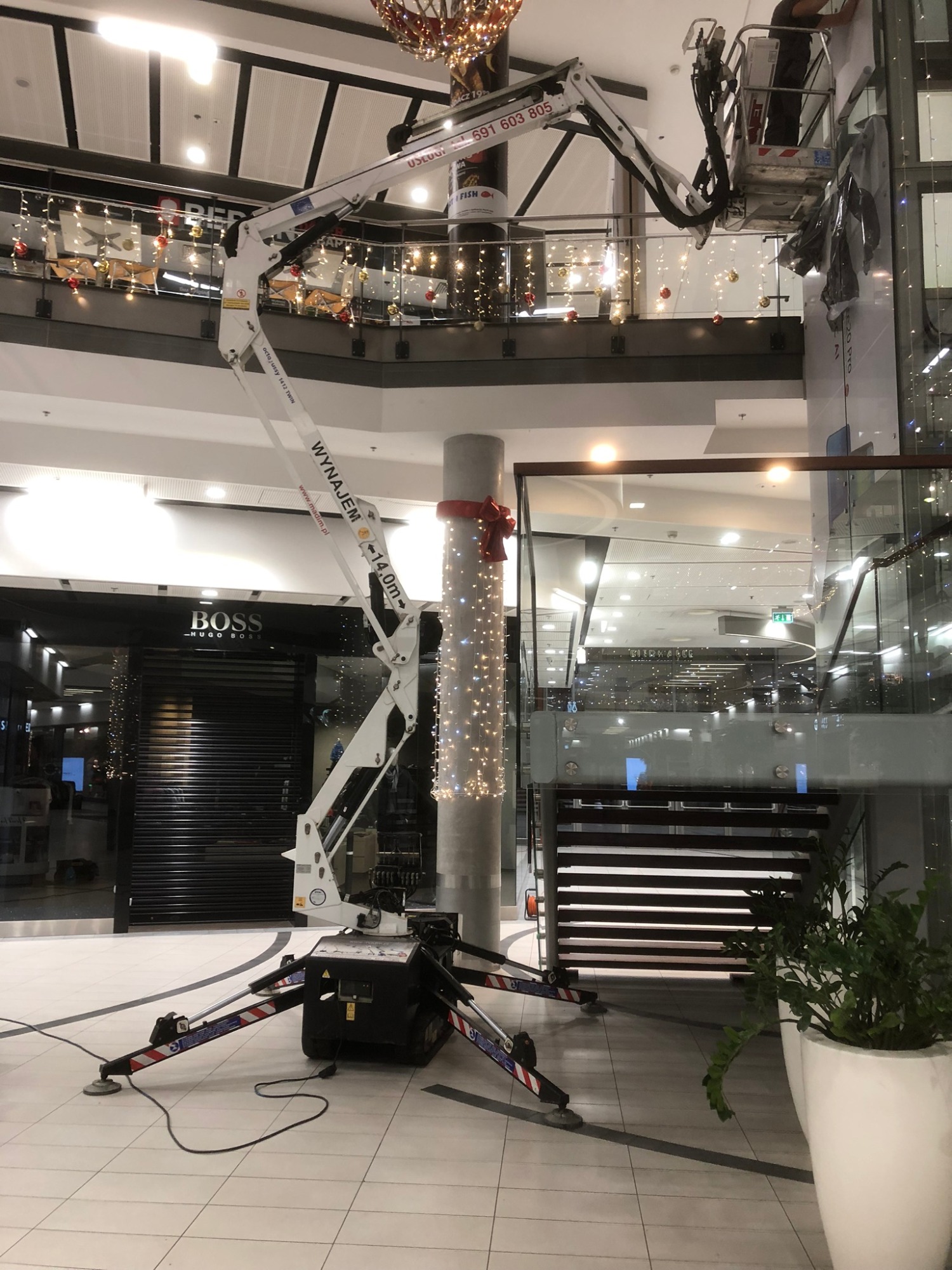 Wysięgnik na podwoziu ustawiony wewnątrz centrum handlowego, dekorowanie kolumny świątecznymi lampkami przez pracownika w koszu, widoczny sklep marki BOSS w tle.