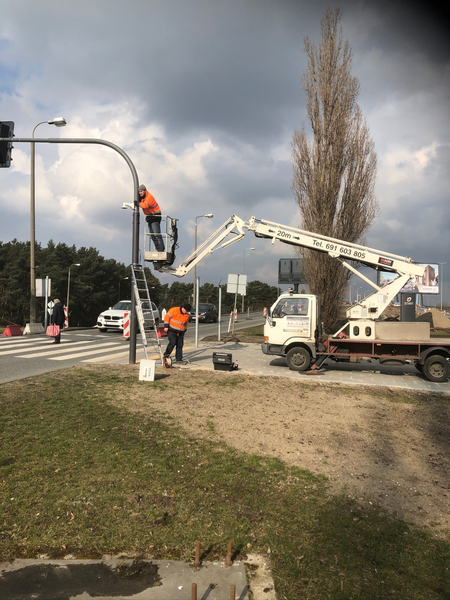 Pracownicy w pomarańczowych kamizelkach montują kamerę na słupie sygnalizacji świetlnej przy użyciu podnośnika koszowego i drabiny, na tle pochmurnego nieba i ulicy z przechodniami.