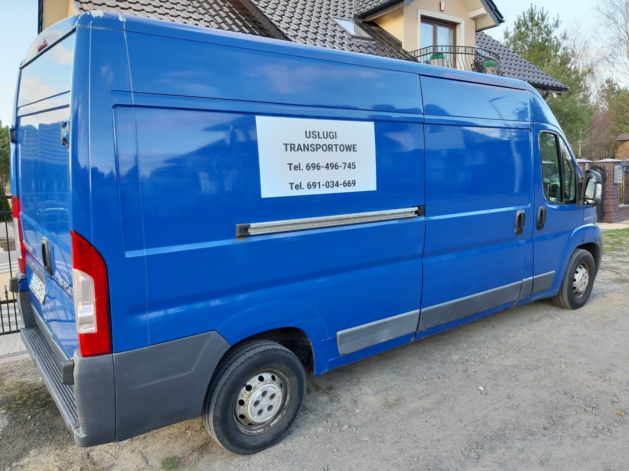 Niebieski samochód dostawczy z naklejoną kartką 'Usługi Transportowe' i numerami telefonów, zaparkowany na żwirowej drodze przed domem.