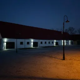 Oświetlony nocą budynek z czerwonym dachem i automatycznymi bramami garażowymi, oświetlony lampami ulicznymi i reflektorami nad bramami.