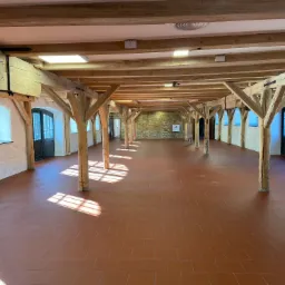 Przestronne, puste wnętrze z drewnianą konstrukcją stropu, terakotową podłogą i promieniami słońca wpadającymi przez okna.