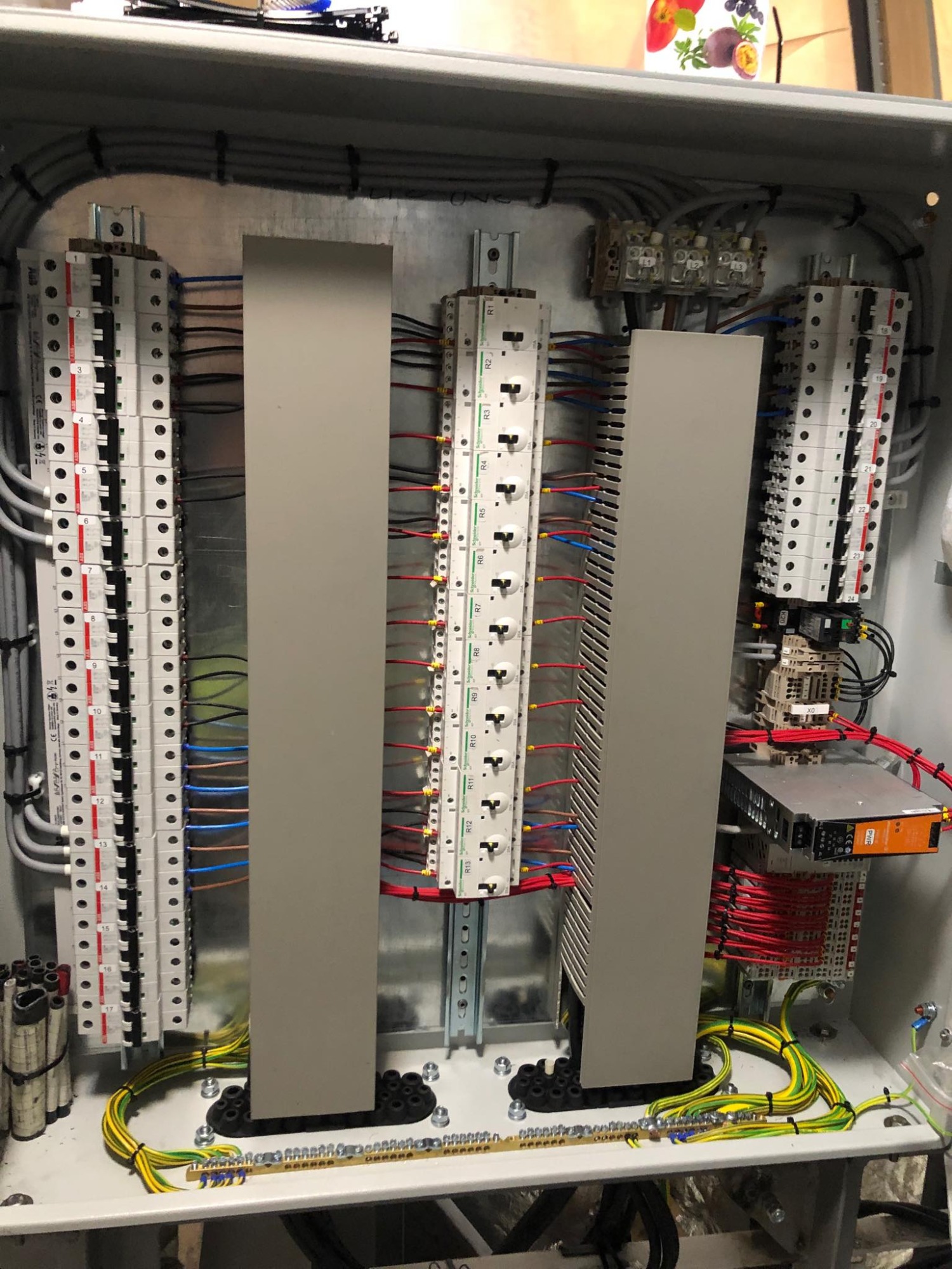 Otwarta skrzynka elektryczna z rozdzielnicą, licznymi przewodami w kolorach szarym, czerwonym, niebieskim i żółto-zielonym, oraz elementami montażowymi, widoczne złącza i bezpieczniki.