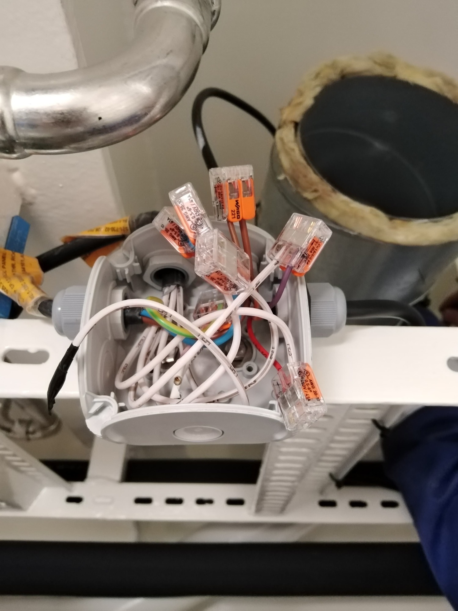 Otwarta, okrągła puszka elektryczna z widocznymi przewodami połączonymi złączkami WAGO, zamontowana pod sufitem z widoczną instalacją wentylacyjną w tle.