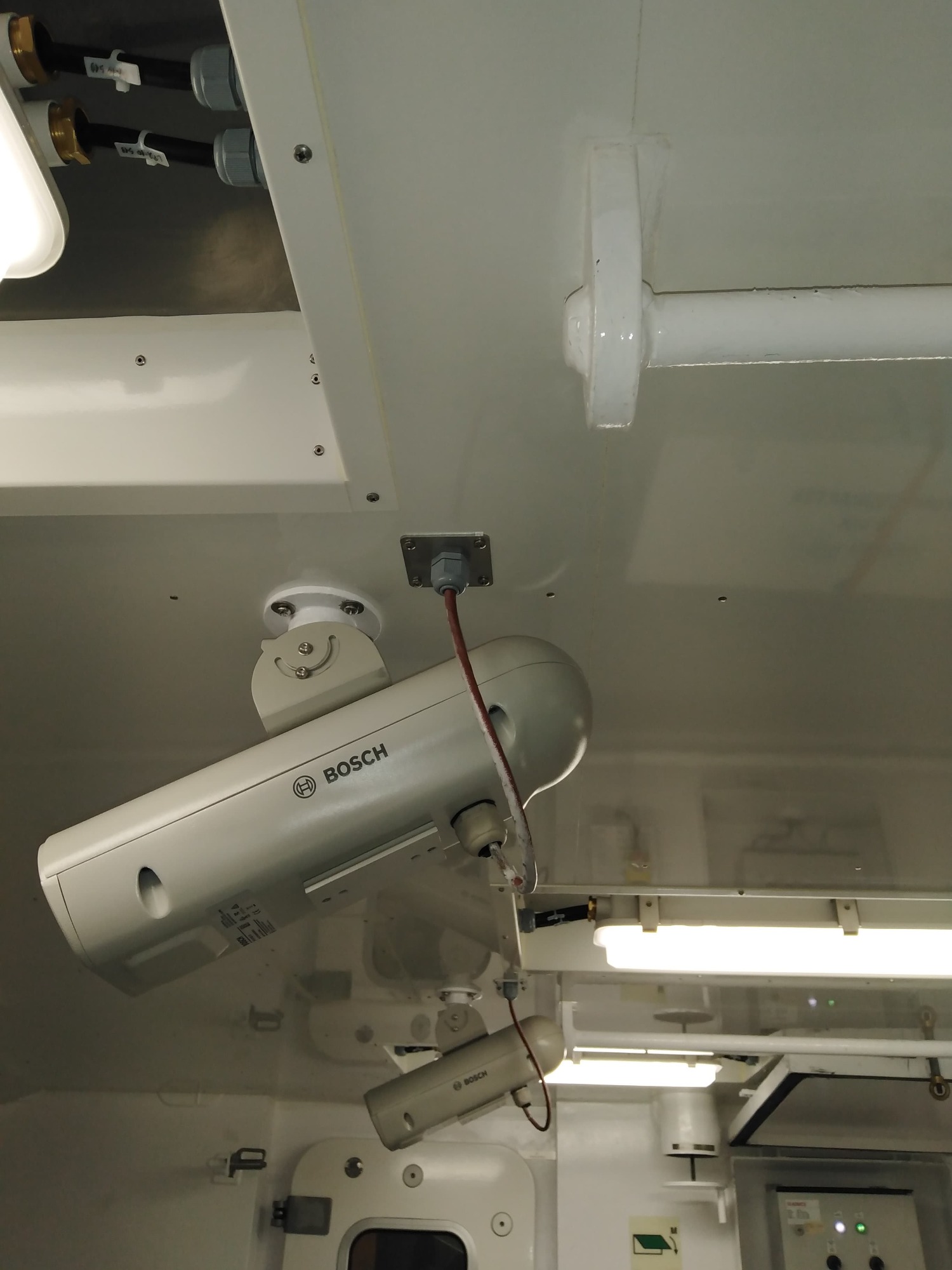 Zamontowane pod sufitem białe kamery przemysłowe marki Bosch z widocznymi przewodami i elementami mocującymi, w tle oświetlenie i elementy instalacji.
