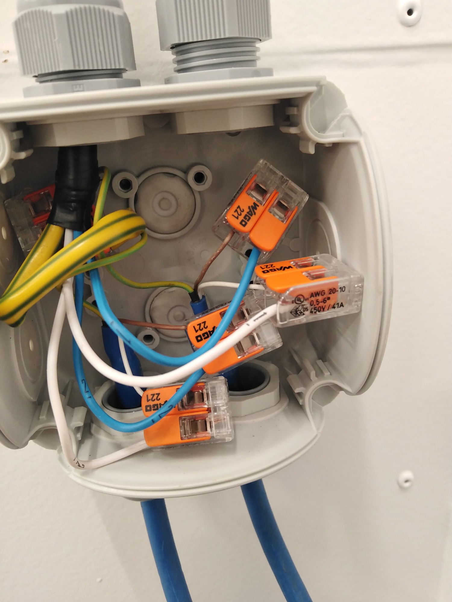 Szczegółowe ujęcie wnętrza szarej puszki elektrycznej z widocznymi przewodami w różnych kolorach (żółto-zielony, niebieski, biały, brązowy) połączonymi za pomocą pomarańczowych złączek WAGO 221...