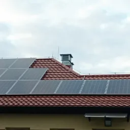 Panele fotowoltaiczne na dachu pokrytym czerwoną dachówką, widoczny komin z metalową osłoną i instalacja odgromowa, pochmurne niebo w tle.