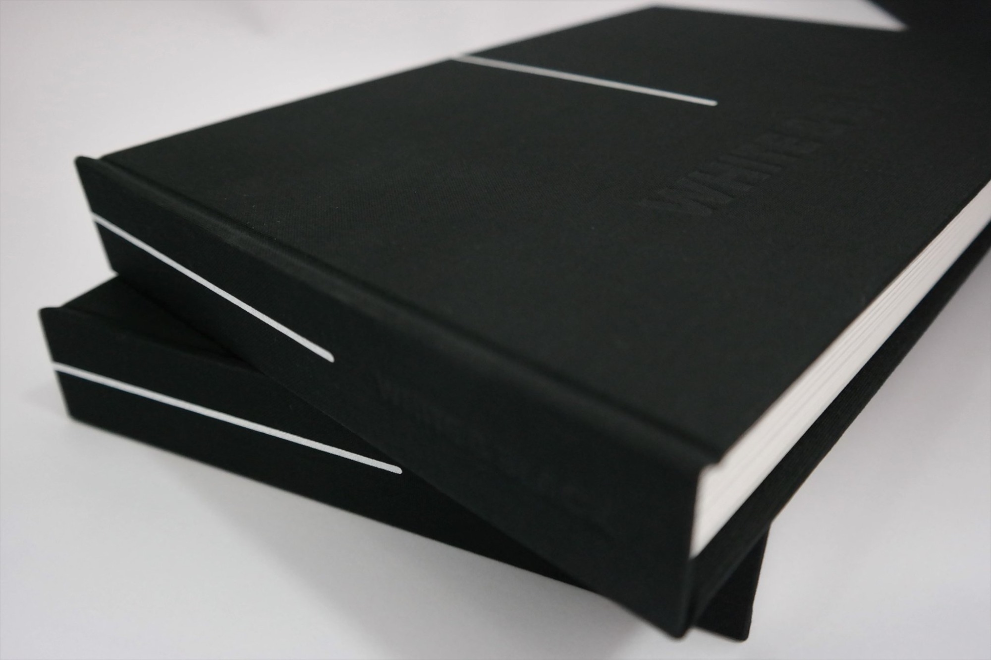 Dwa czarne, twarde oprawione albumy, jeden na drugim, z wytłoczonym napisem i białymi liniami dekoracyjnymi na okładce.
