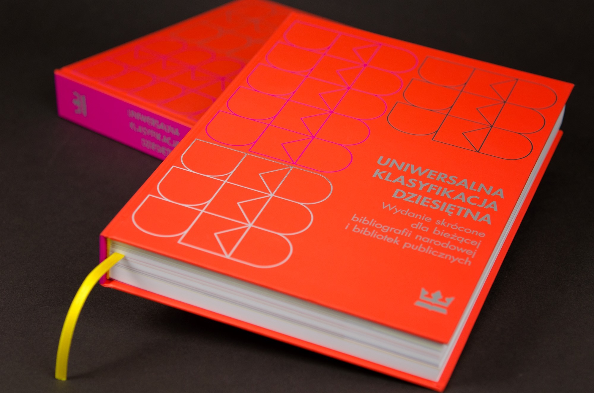 Dwa identyczne, pomarańczowe tomy książki w twardej oprawie z abstrakcyjnym wzorem i srebrnymi napisami, jeden z żółtą zakładką, leżące na ciemnym tle.