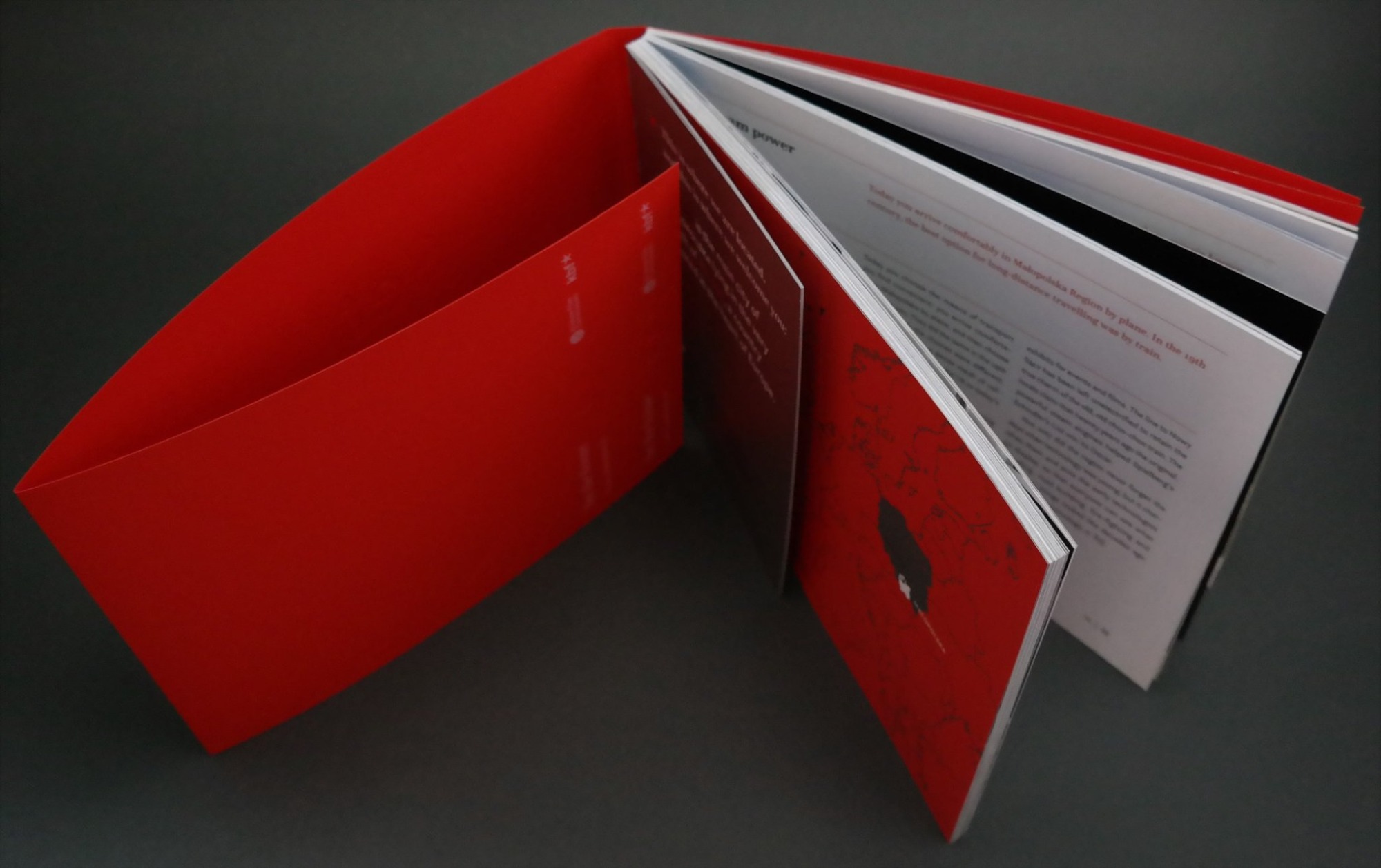 Otwarty, czerwony folder firmowy z widoczną zawartością: tekstem i grafiką w kolorach czerwieni, bieli i czerni, leżący na ciemnoszarym tle.