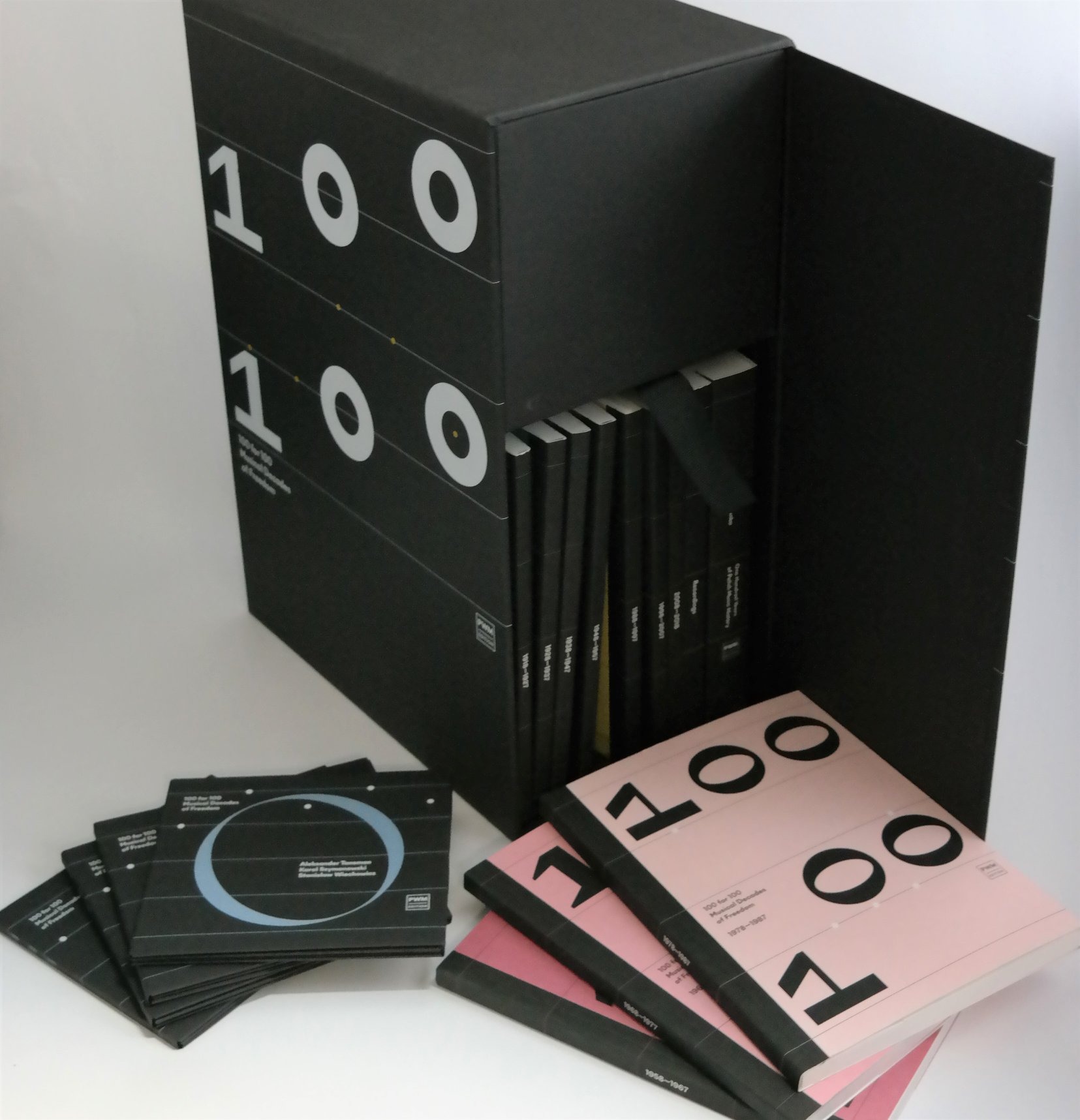 Zestaw katalogów w czarnym pudełku z napisem '100', obok leżą dodatkowe katalogi w kolorach czarnym i różowym.