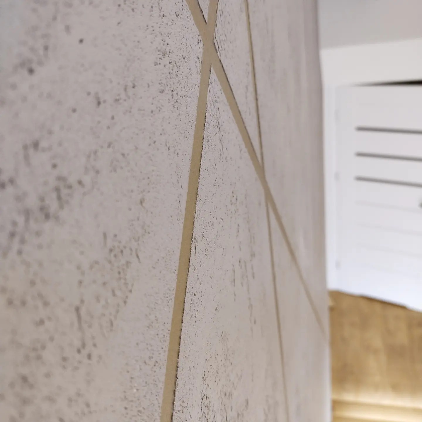 Dekoracyjna ściana z efektem betonu i geometrycznym wzorem w kolorze złamanej bieli, widoczna z bliskiej perspektywy, z elementami drzwi w tle.