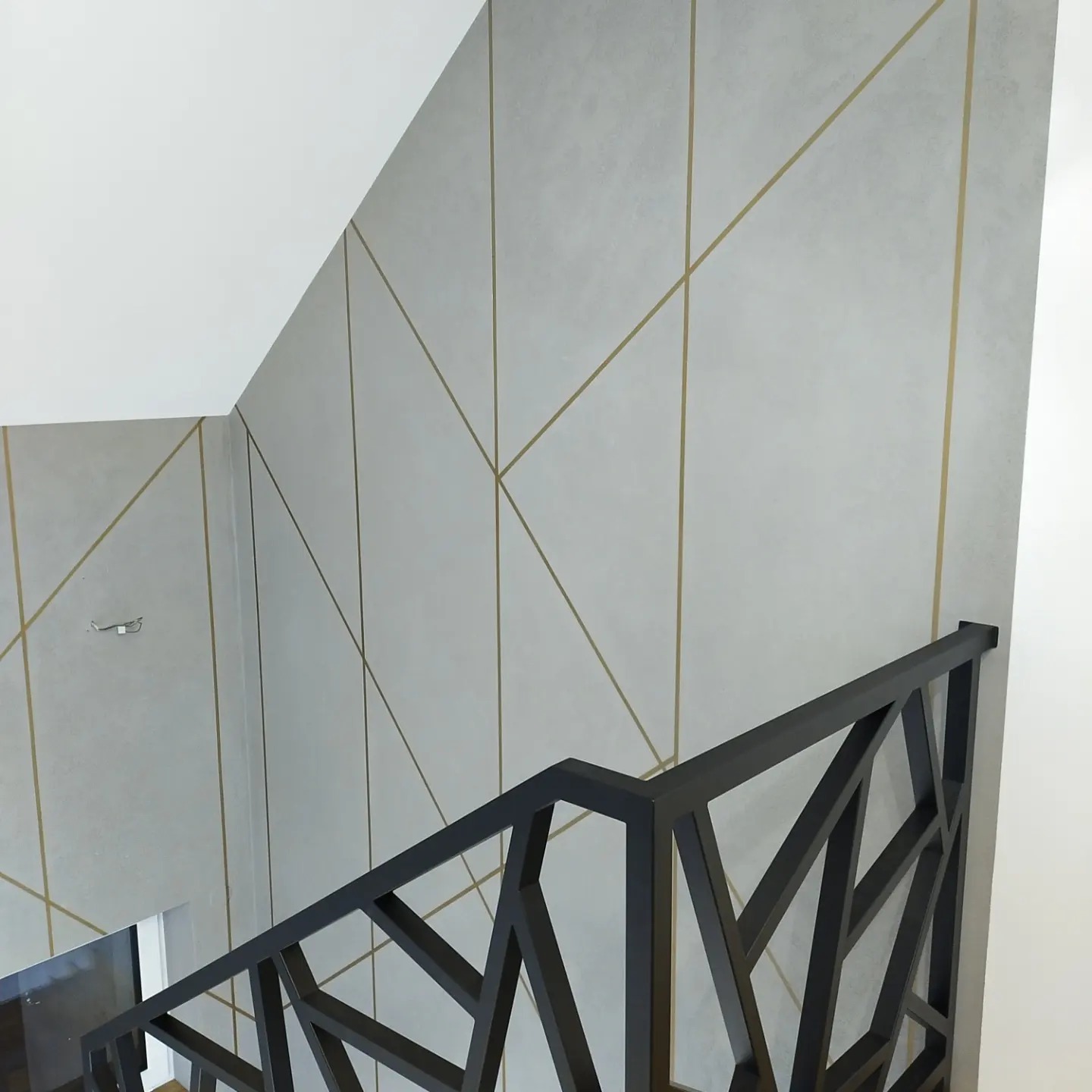 Wnętrze klatki schodowej z geometrycznym wzorem z cienkich złotych linii na szarej ścianie i nowoczesną, czarną balustradą o ażurowej konstrukcji.