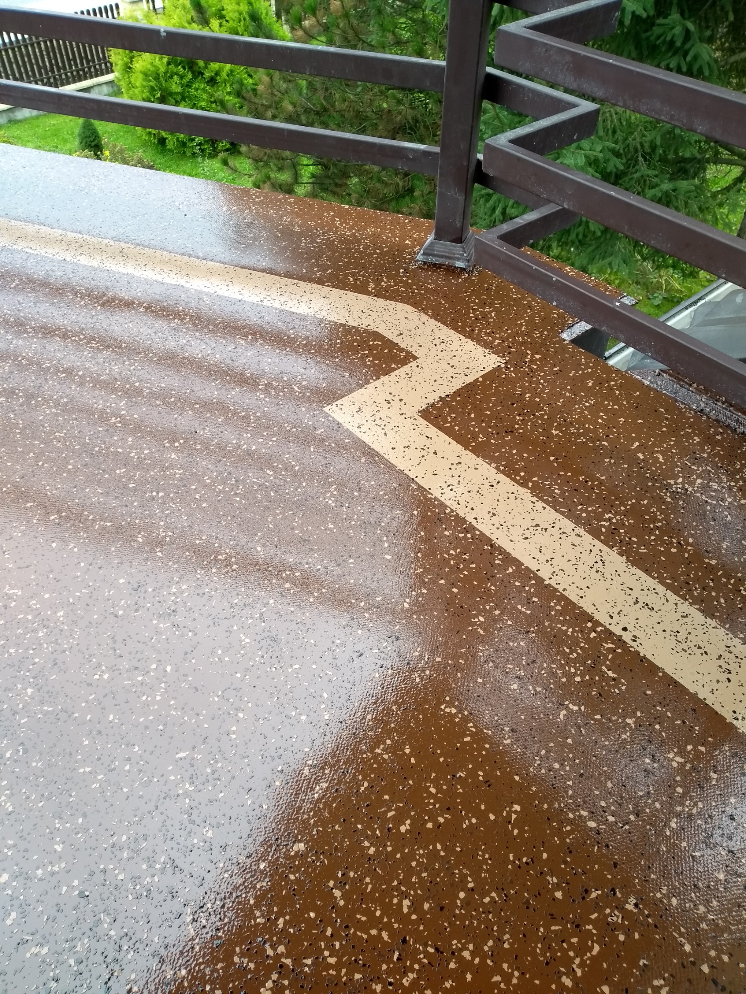 Błyszcząca, brązowa posadzka żywiczna na balkonie z dekoracyjnym, jasnym zygzakiem; widoczna metalowa balustrada i zieleń w tle.