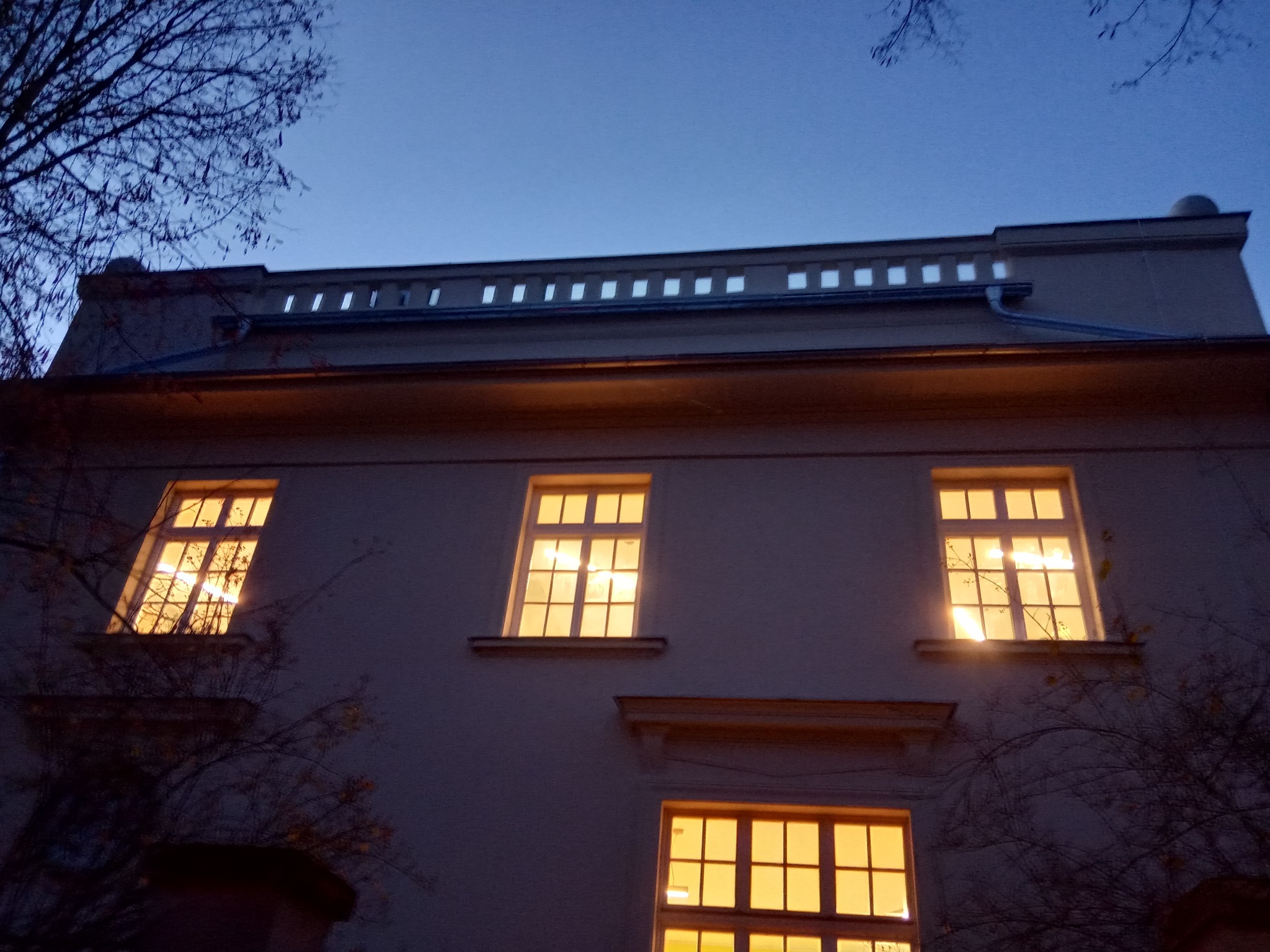 Elewacja budynku z oknami oświetlonymi od wewnątrz, widoczny dach z rynnami i dekoracyjnym elementem w górnej części, sfotografowane o zmierzchu.