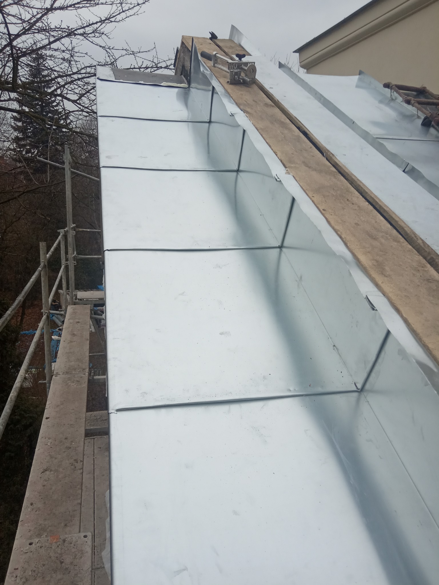 Widok z góry na fragment dachu pokrytego błyszczącymi, metalowymi panelami, z drewnianą deską i narzędziem dekarskim na górze, obok rusztowania i drzew bez liści, przy pochmurnym niebie.