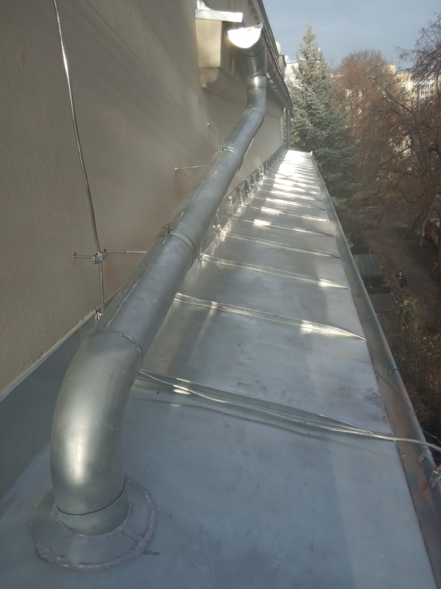 Nowe metalowe orynnowanie i blaszany dach z widocznymi elementami mocującymi do ściany budynku oraz drzewami w tle.