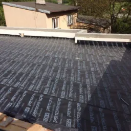 Radzymin: Czarna membrana hydroizolacyjna Tack-R na płaskim dachu, z widocznymi elementami betonowej attyki i fragmentami drewnianej konstrukcji, w tle budynek mieszkalny.