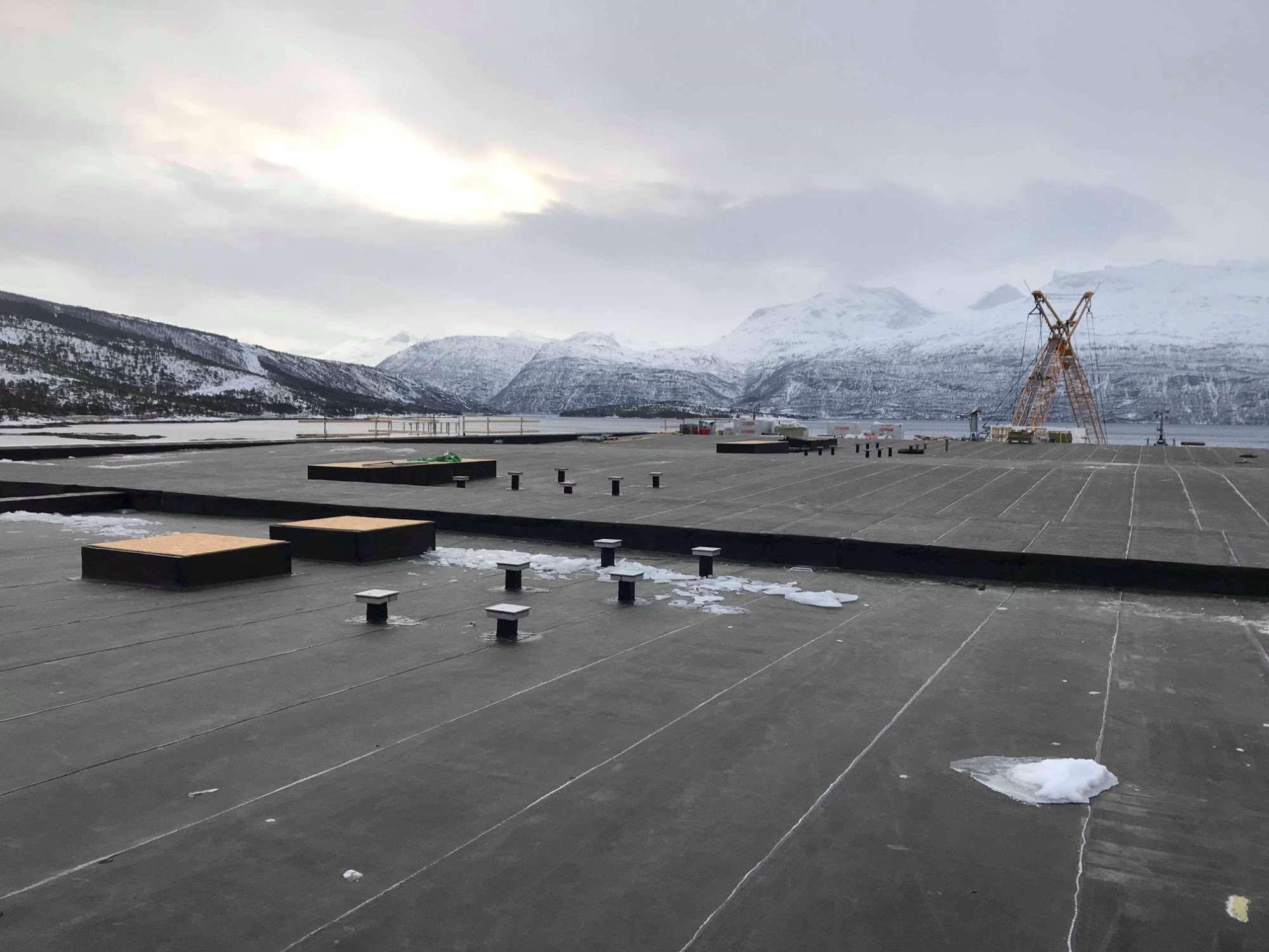 Widok na rozległy, płaski dach pokryty czarną membraną Derbigum Norway, z widocznymi odpowietrznikami i niewielkimi plamami śniegu, na tle zimowego krajobrazu z górami i dźwigiem.