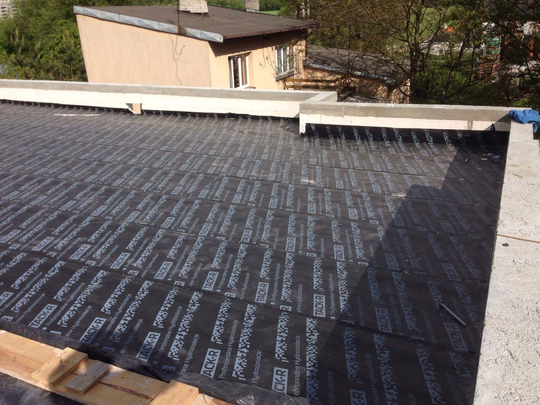 Radzymin: Czarna membrana hydroizolacyjna Tack-R na płaskim dachu, z widocznymi elementami betonowej attyki i fragmentami drewnianej konstrukcji, w tle budynek mieszkalny.