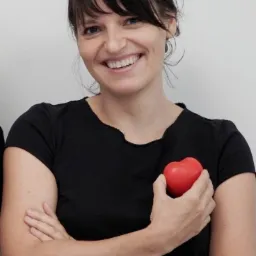 Uśmiechnięta kobieta z Poznania w czarnej koszulce trzymająca czerwoną gumową zabawkę w kształcie serca przy piersi.