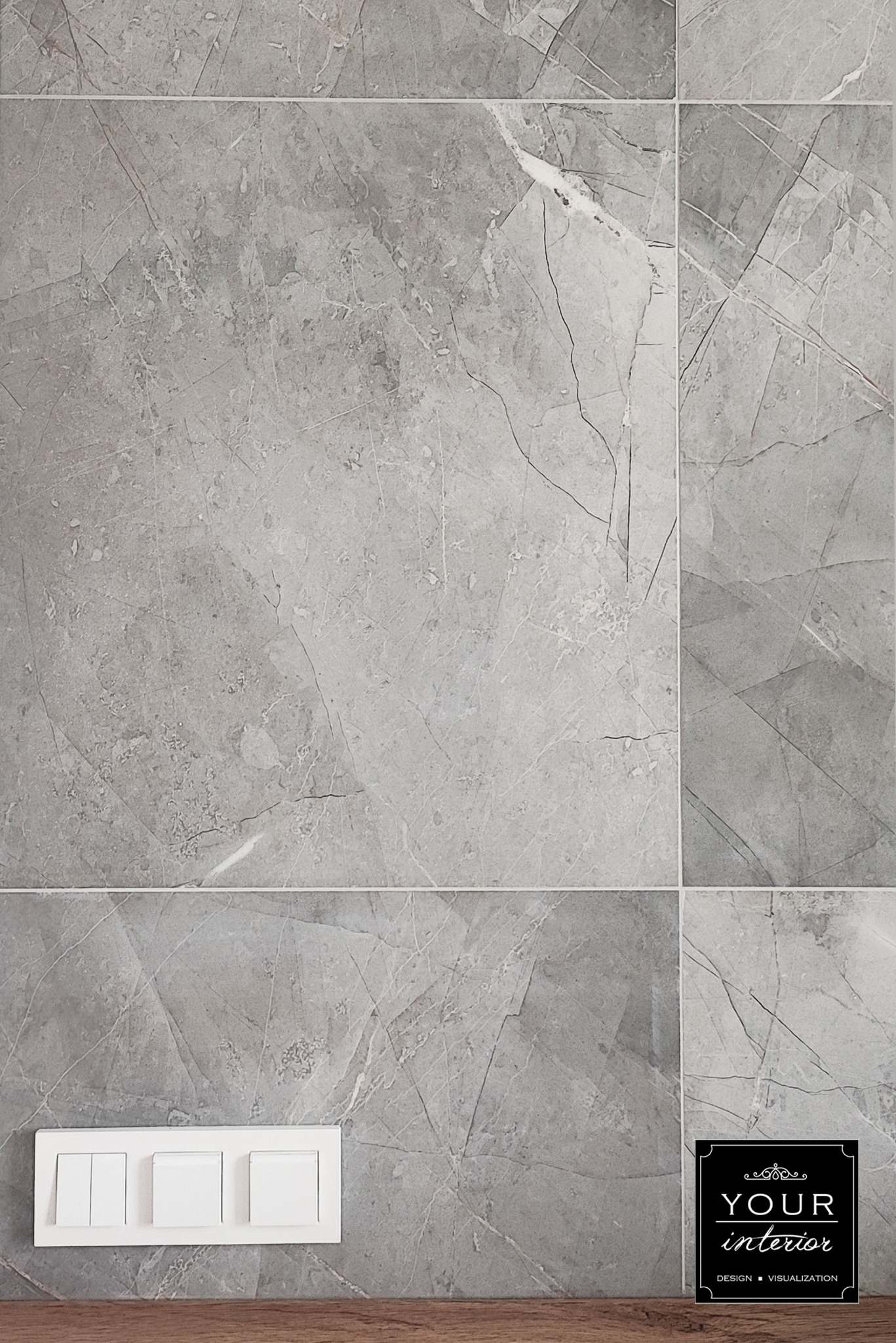 Ściana wyłożona szarymi płytkami imitującymi marmur, z widocznym panelem z trzema włącznikami światła i fragmentem drewnianej podłogi u dołu. W prawym dolnym rogu widoczne logo firmy.