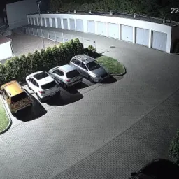Nocny widok z kamery monitoringu na parking z kilkoma samochodami, garażami i zielenią, data i godzina w prawym górnym rogu.