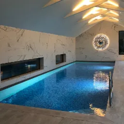 Kryty basen w nowoczesnym stylu z marmurowymi ścianami, oświetleniem sufitowym w kształcie strzałek i dekoracyjnym okrągłym elementem ściennym, widok na taflę wody z delikatnymi refleksami.