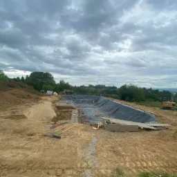 Przygotowanie wykopu pod zbiornik wodny z rozłożoną czarną folią hydroizolacyjną, widoczne narzędzia budowlane i koparka CAT w tle.