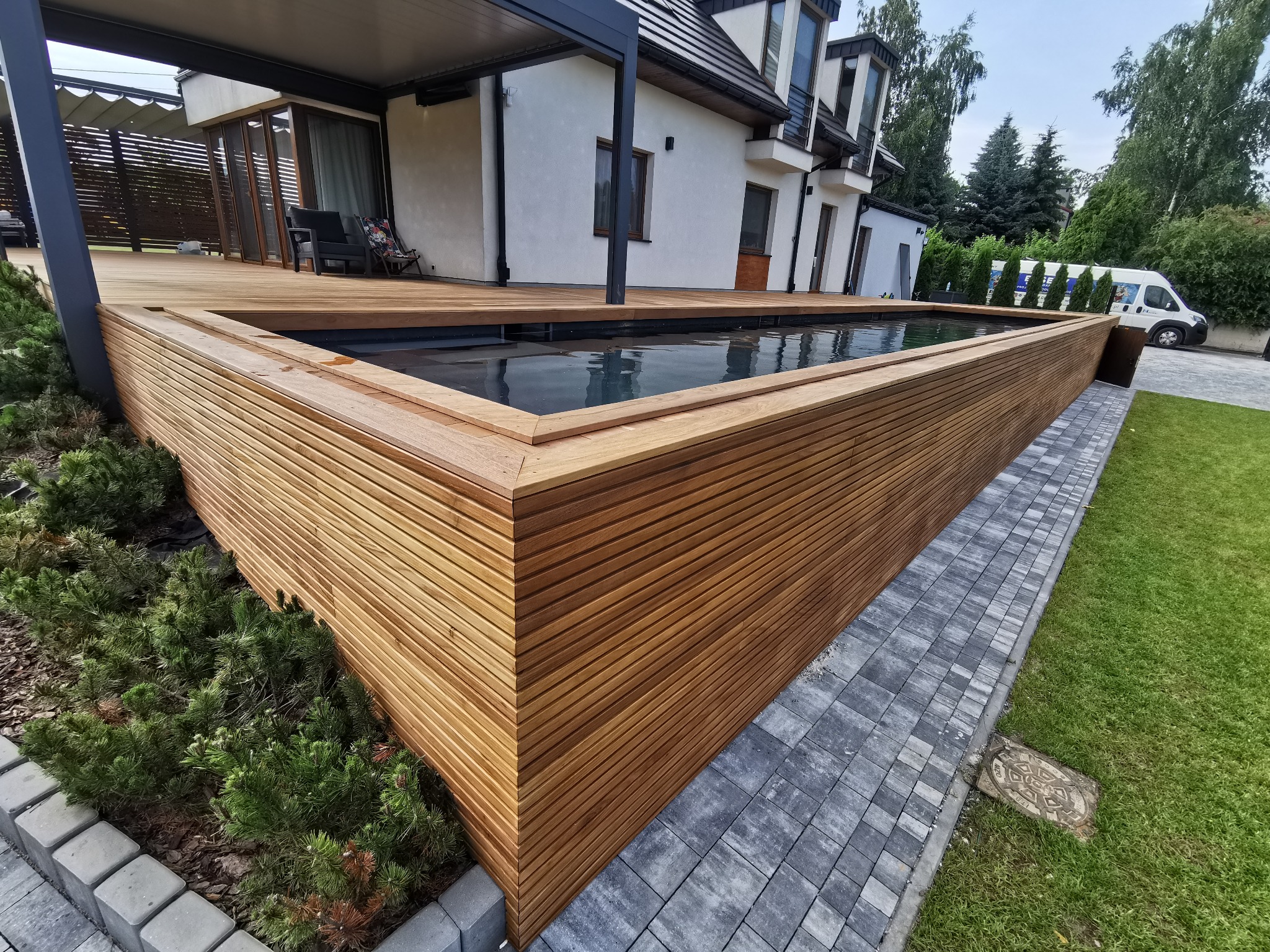 Nowoczesny basen z obudową z drewna, płynnie przechodzący w taras, z widocznym fragmentem domu w tle i starannie ułożoną kostką brukową.