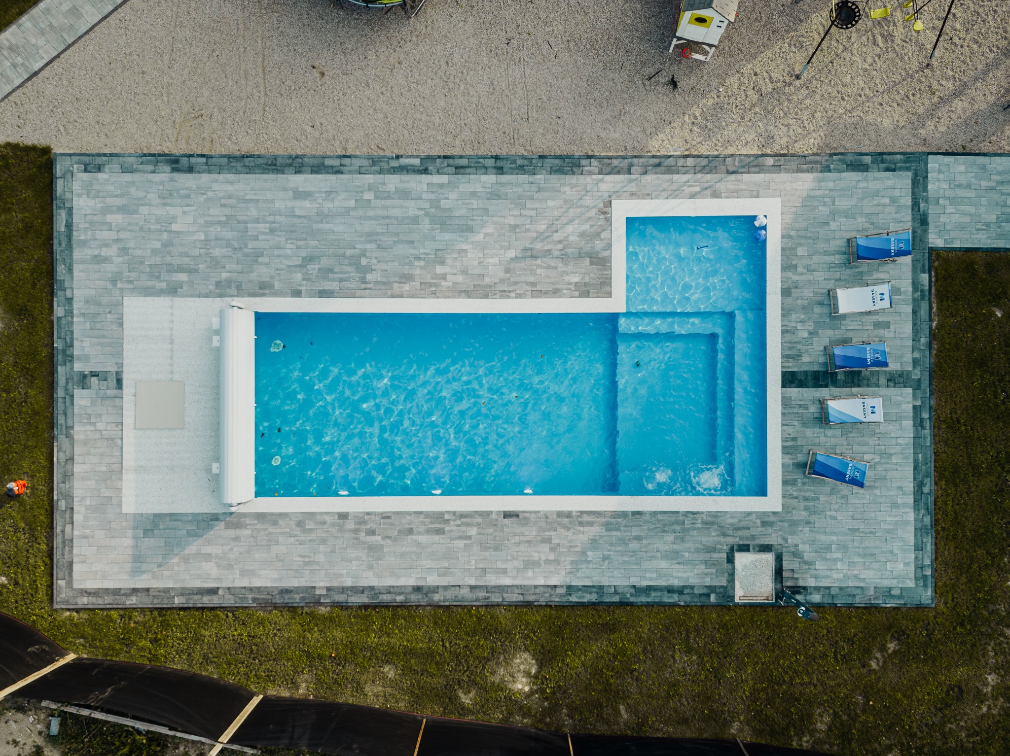 Widok z góry na prostokątny basen z brodzikiem, otoczony szarą kostką brukową, obok plac zabaw dla dzieci i leżaki z logo firmy.