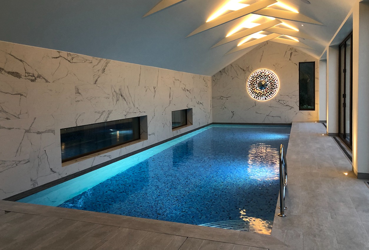 Kryty basen w nowoczesnym stylu z marmurowymi ścianami, oświetleniem sufitowym w kształcie strzałek i dekoracyjnym okrągłym elementem ściennym, widok na taflę wody z delikatnymi refleksami.