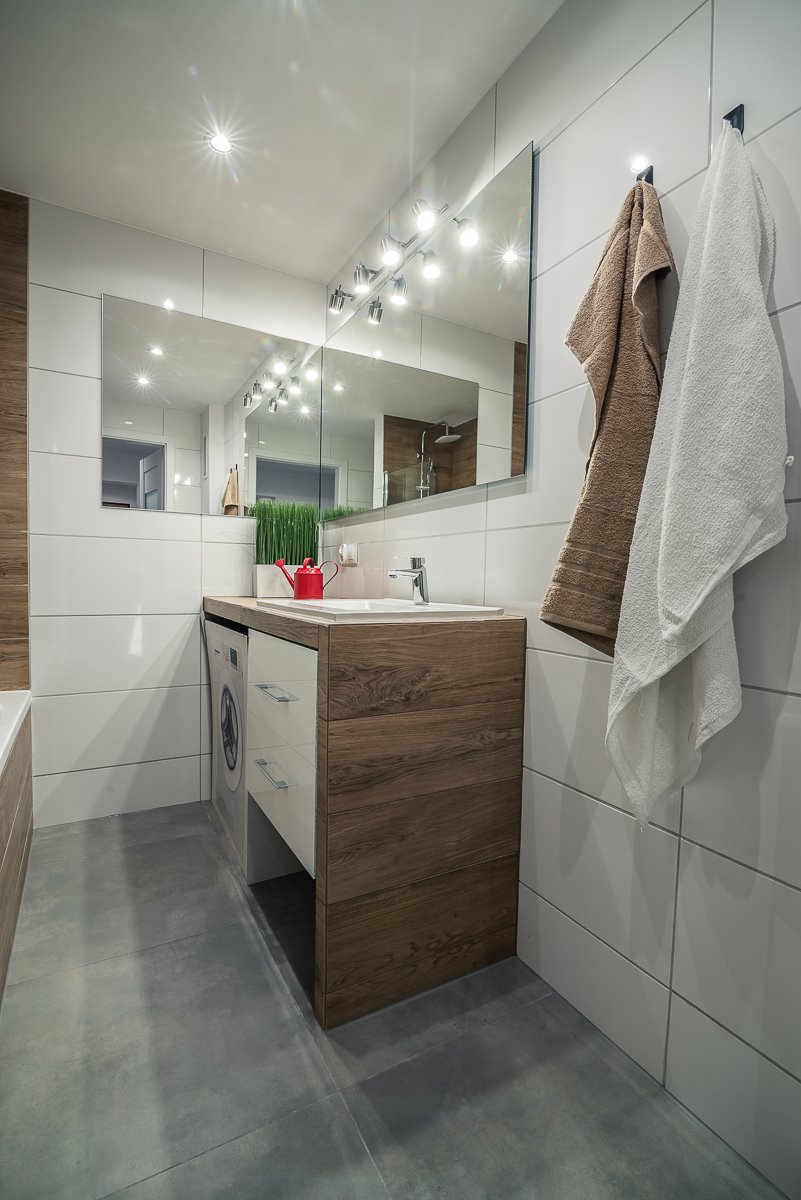 Nowoczesna łazienka z drewnianą szafką pod umywalką, wbudowaną pralką, dużym lustrem z oświetleniem punktowym i dwoma ręcznikami na białych płytkach.
