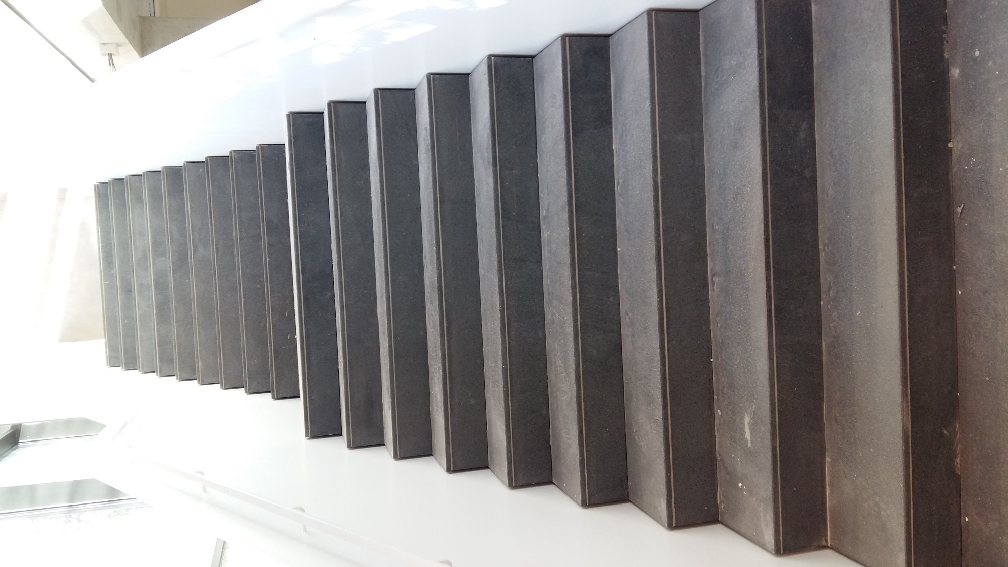 Szereg pionowych, ciemnych listew z kamienia lub betonu tworzących dekoracyjny element elewacji budynku, widoczne z dołu pod kątem.
