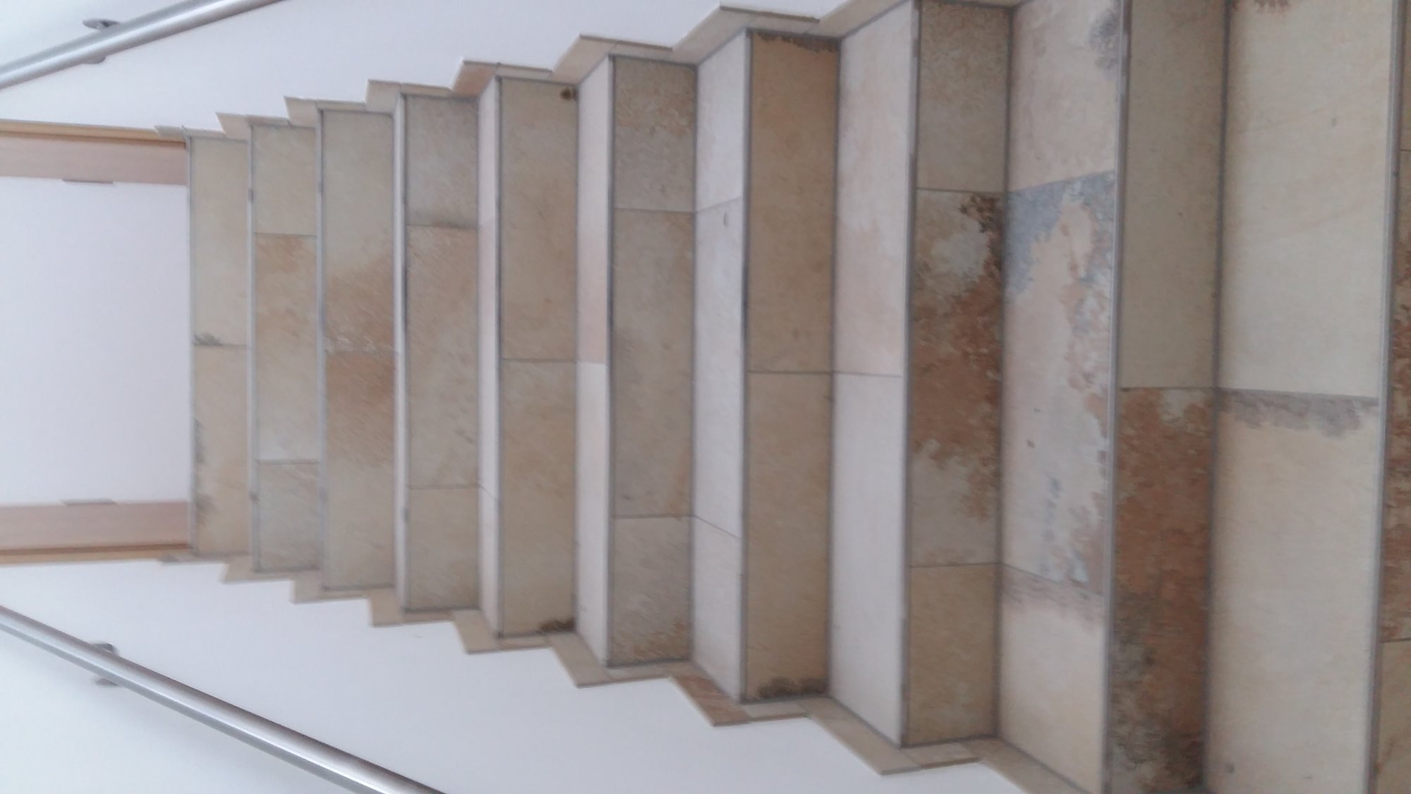 Widok z góry na wyłożone płytkami schody z metalową poręczą, oświetlone światłem dziennym, z widocznymi szarymi fugami i różnymi odcieniami beżu i brązu na płytkach.