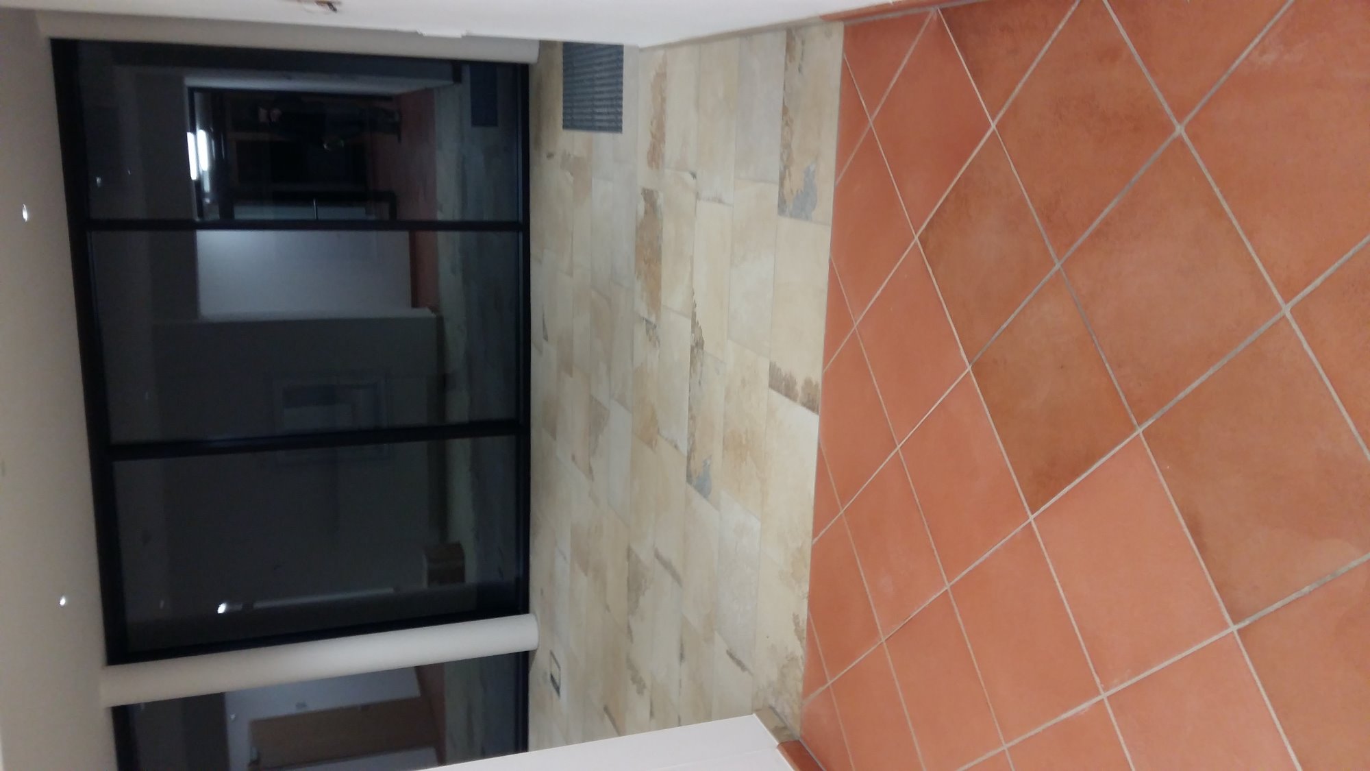 Wnętrze z terakotową podłogą ułożoną w karo, beżową ścianą z efektem kamienia i widocznym fragmentem okna z czarną ramą, odbijającego przestrzeń wewnątrz budynku.