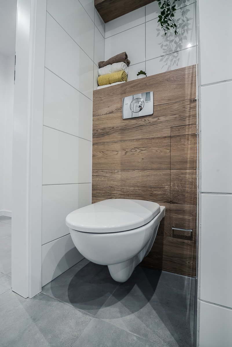 Nowoczesna, podwieszana toaleta z chromowanym przyciskiem spłukiwania na tle drewnopodobnych paneli, uzupełniona o białe płytki i szare płytki podłogowe w minimalistycznej łazience.