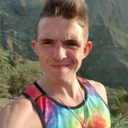 Mężczyzna w koszulce tie-dye robi selfie na tle górzystego krajobrazu z palmami, prawdopodobnie podczas treningu biegowego na świeżym powietrzu.