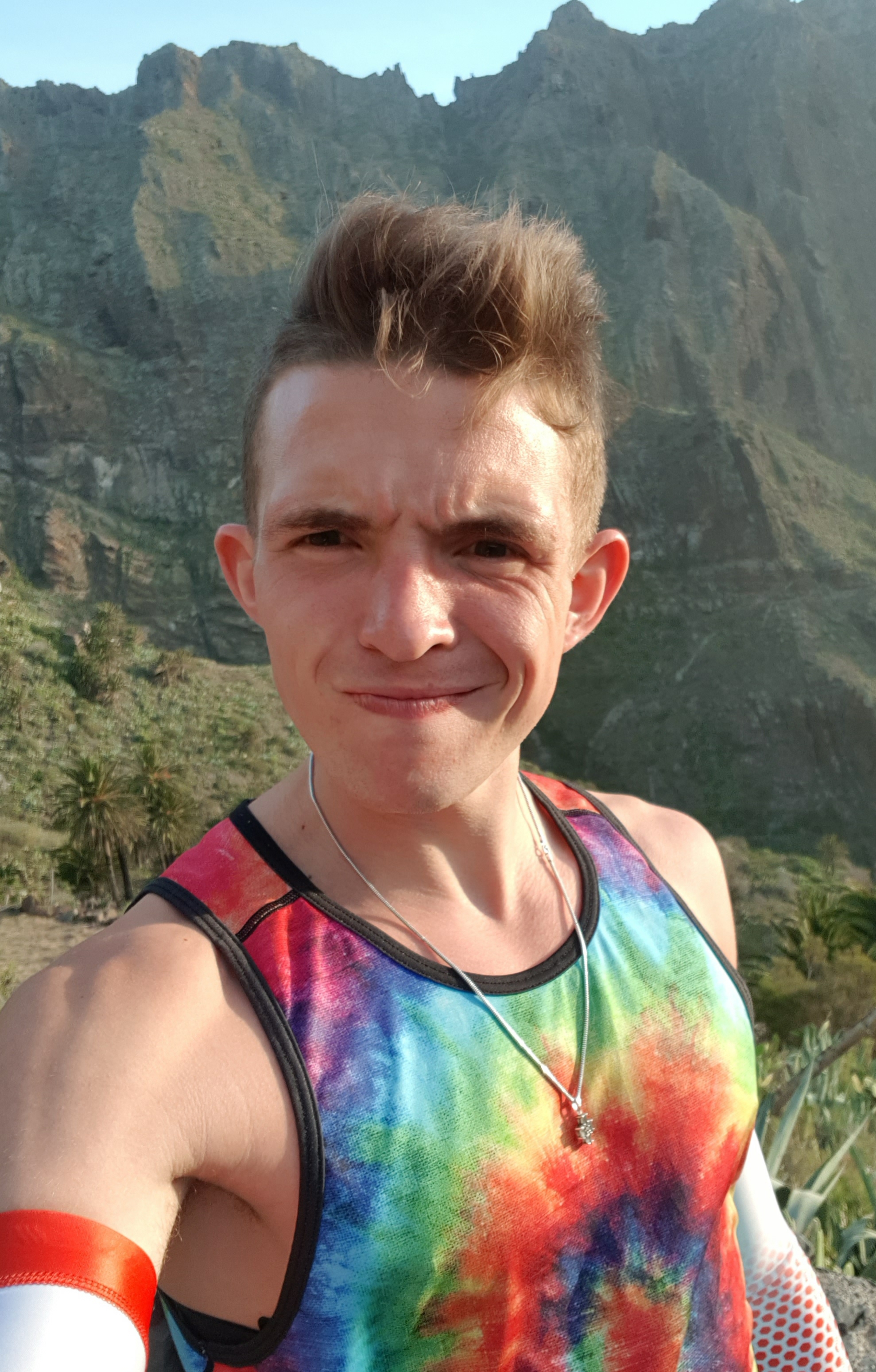 Mężczyzna w koszulce tie-dye robi selfie na tle górzystego krajobrazu z palmami, prawdopodobnie podczas treningu biegowego na świeżym powietrzu.