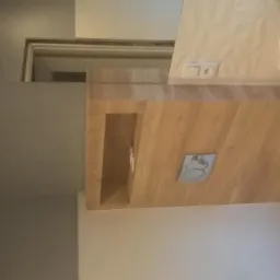 Nowoczesna łazienka z drewnianą zabudową, wiszącą toaletą i umywalką nablatową. Widoczny fragment lustra i dekoracyjna płytka na ścianie.