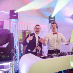 Dwóch mężczyzn w strojach galowych prezentuje sprzęt DJ-ski i oświetlenie sceniczne podczas imprezy, jeden z mikrofonem, drugi z laptopem Apple.