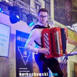 Mężczyzna w okularach gra na czerwonym akordeonie marki Roland na scenie z oświetleniem, widoczne banery reklamowe i mikrofon.