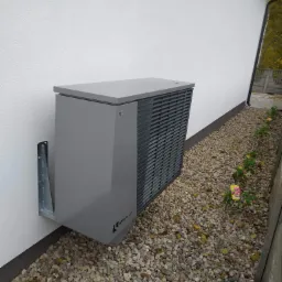 Szara jednostka zewnętrzna z logo Alpha Innotec, zamontowana na białej ścianie budynku, widoczna z boku, z elementami montażowymi i kamiennym podłożem.